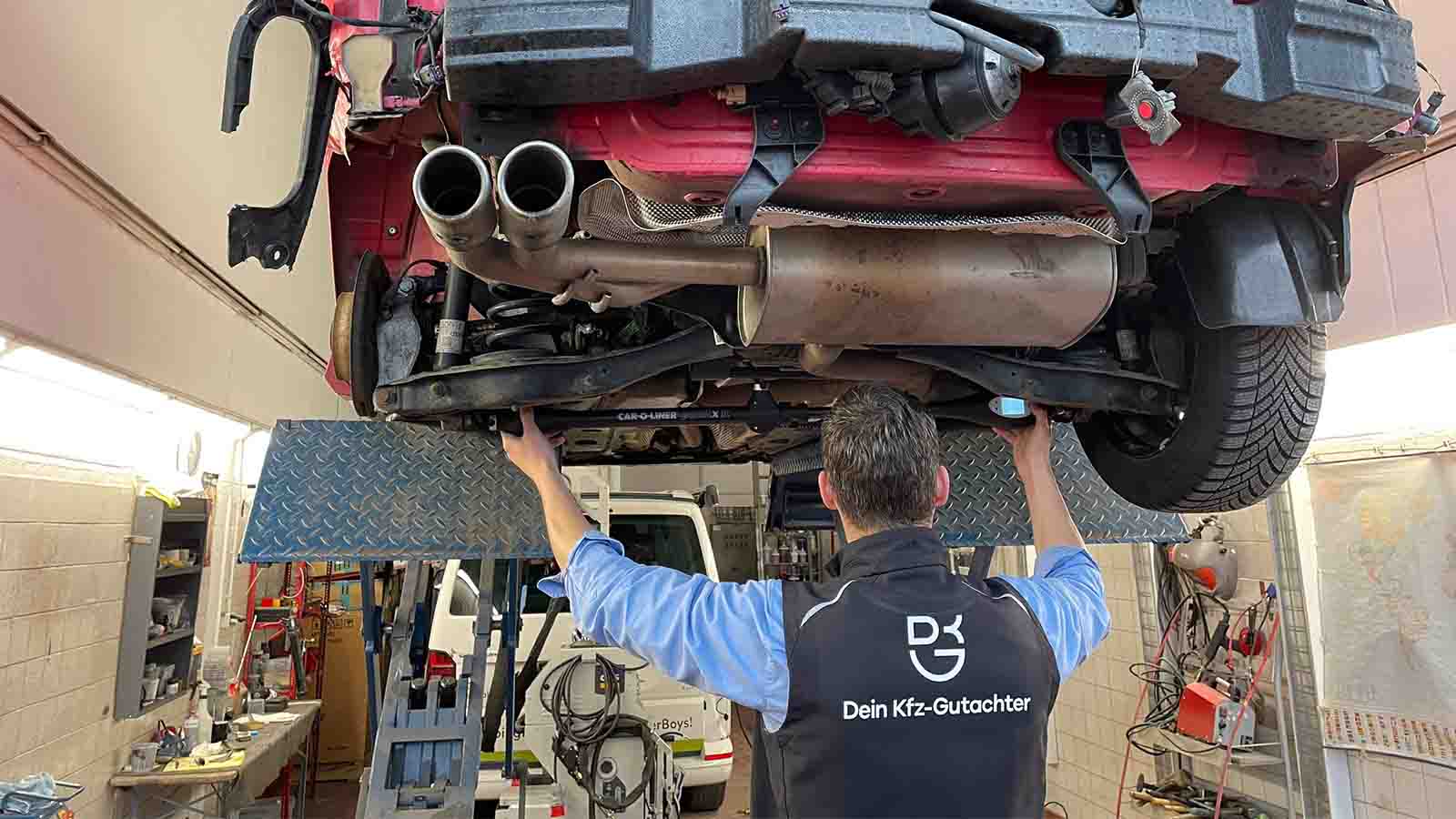 Sebastian Wyczisk von hinten in Arbeitsweste „Dein Kfz-Gutachter“, er setzt unter der Hebebühne eine Messbrücke am Unterboden eines Unfallautos an